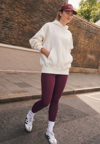 Weißes Kapuzen-Sweatshirt mit Taschen, burgunderrote Leggings und weiße Turnschuhe mit grünen Akzenten. Dazu eine burgunderrote Mütze, auf einer Straße getragen.