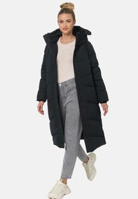 Long black puffer coat with a zip and button closure, featuring a hood and quilted texture. Paired with light gray pants and white sneakers.