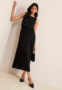 Schwarzes Maxikleid mit ärmellosem Design und gerafftem Taillienbereich. Der Stoff ist glatt; kombiniert mit schwarzen spitzen High Heels und einer kleinen Handtasche.