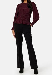 Mujer vestida con una blusa burdeos de manga larga y fruncida, pantalones negros rectos y botas negras de tobillo, de pie con una mano en el bolsillo.