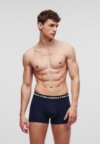 Férfi modell, göndör hajjal, sötétkék Karl Lagerfeld boxer alsót viselve, egy egyszínű világosszürke háttér előtt áll.