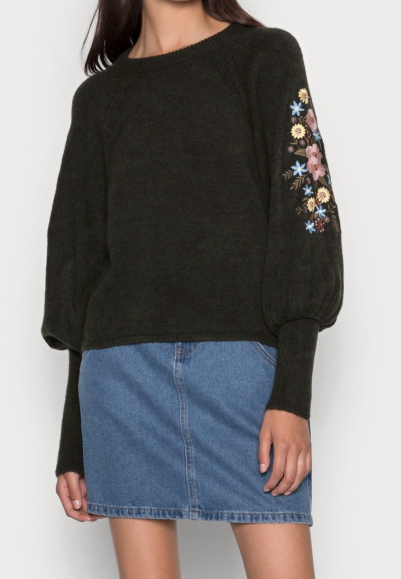 Pull en tricot vert foncé avec des manches oversized, orné d'une broderie florale rose et bleue sur le bras gauche, accompagné d'une jupe courte en denim.