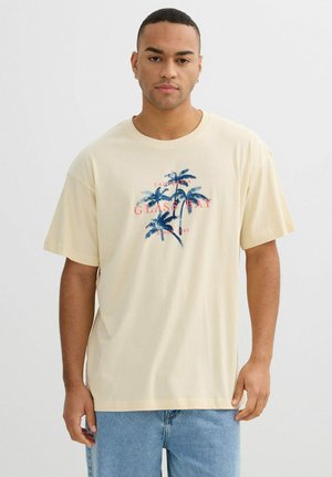 Joven con una camiseta beige con palmeras azules y texto rojo que dice "California Glass Bay Since 1999", combinada con vaqueros azul claro.