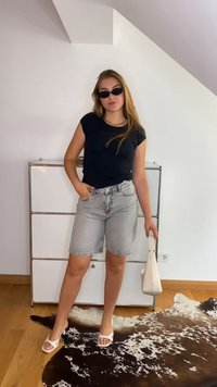 Svart åtsittande t-shirt, grå denimshorts och vita sandaler. Håller en liten vit handväska. Står på en getskinnsmatta, mot en enkel bakgrund.