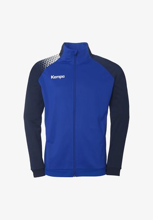 Blå atletisk jakke med marineblå accenter, fuld lynlås foran, høj krave og geometrisk mønster på den ene skulder. Har "Kempa" logo på brystet.