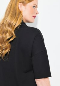 Top negro de manga corta con un diseño clásico, textura suave y un escote alto, con cabello largo y ondulado visible en el lado izquierdo.