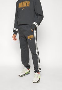 Nike-huppari ja verkkarit tummanharmaana, valkoisilla raidoilla, joissa on näkyvä kultainen "NIKE"-brändäys. Kengät vihreillä yksityiskohdilla.