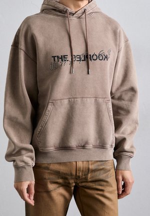 Osoba ubrana w wyblakłą brązową bluzę z kapturem z kieszenią kangurzą i zdeformowanym czarnym napisem na klatce piersiowej, w połączeniu z brązowymi spodniami.