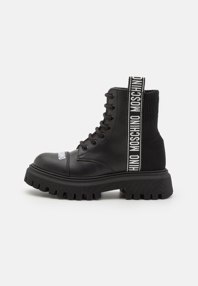 Botte de combat en cuir noir avec semelle épaisse à relief, laçage à l'avant, panneau en maille au niveau de la cheville, et sangle blanche marquée "Moschino" sur le côté et le bout de la chaussure.