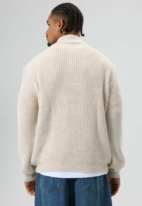 Man met gevlochten haar, gekleed in een beige gebreide trui en blauwe spijkerbroek, met de rug naar de camera tegen een effen grijze achtergrond.