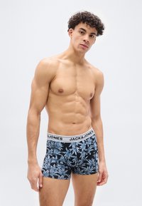 Νέος άνδρας με σγουρά μαλλιά που φοράει μαύρα και μπλε σλιπ με σχέδιο Jack & Jones, στέκεται μπροστά σε απλό ανοιχτόχρωμο φόντο.