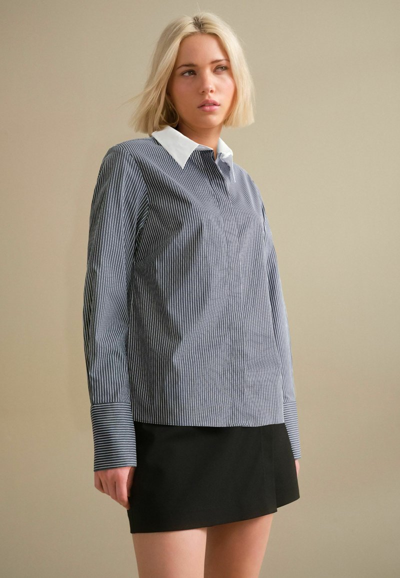 Striped button-up shirt in navy and white with a white collar, long sleeves, and a straight cut. Paired with a black skirt.