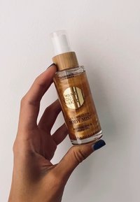 Genomskinlig glasflaska med ett bambulock, som innehåller gyllene vätska. Etiketten läser "Golden Glow Body Mist" och framhäver fuktgivande och glittrande egenskaper.