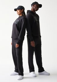 Μαύρα φούτερ με κουκούλα και τυπωμένα λογότυπα, συνδυασμένα με ταιριαστά παντελόνια τζόγκινγκ. Και οι δύο φορούν μαύρα καπέλα και λευκά αθλητικά παπούτσια, με υφή λείου υφάσματος.