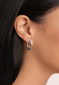 Pendiente de aro de plata con un diseño texturizado que presenta una fila de pequeñas piedras brillantes a lo largo del borde superior. Acabado pulido, forma circular.
