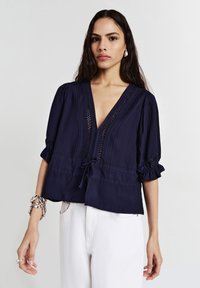 Donna che indossa una blusa blu navy con scollatura a V e dettagli in pizzo, abbinata a pantaloni bianchi, accessoriata con orecchini d'argento, bracciale e anelli.