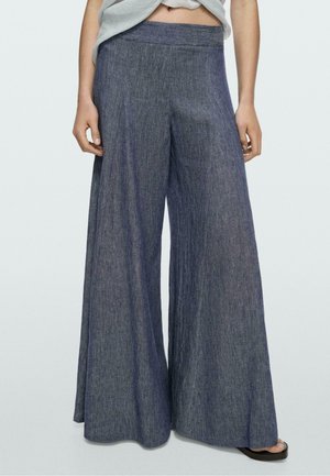 Kobieta w niebieskich dżinsach z wysokim stanem i szerokimi nogawkami, w połączeniu z krótkim jasnoszarym topem i czarnymi klapkami.