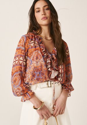 Femme aux longs cheveux noirs, portant un chemisier orange à motifs avec des volants, un pantalon blanc taille haute et un bracelet en or, tenant une sangle beige.