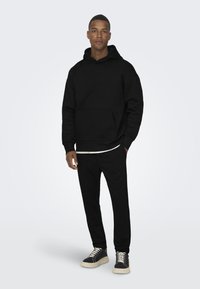 Hanorac negru cu buzunar tip cangur, asortat cu pantaloni negri și teniși negri cu șireturi albe și talpă texturată. Design simplu și coerent.
