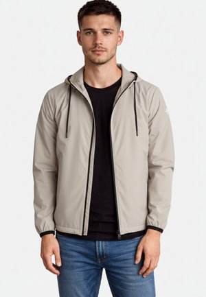 Młody mężczyzna w jasnoszarej kurtce z kapturem na czarnej koszulce i niebieskich dżinsach, stojący na tle jednolitego białego tła.