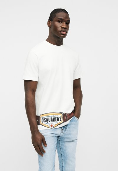 Jeune homme portant un t-shirt blanc uni, un jean bleu clair et une ceinture marron avec une grosse boucle DSQUARED2, debout avec une main dans la poche.
