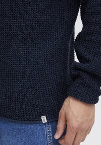 Marineblauer Strickpullover mit strukturiertem Muster, ausgestattet mit gerippten Bündchen und einem Etikett am Saum. Getragen mit Jeans.