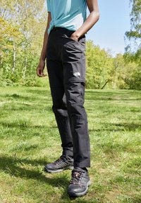 Svarta friluftsbukser har en dragkedjeförsedd cargo-laga och logodetalj, i kombination med svarta vandringsskor med orange accenter, placerat på gräs.