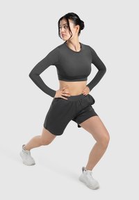 Donkergrijs langemouwen crop top en bijpassende shorts, gemaakt van soepel materiaal. Draag met lichtgekleurde sportschoenen in een lunges houding.