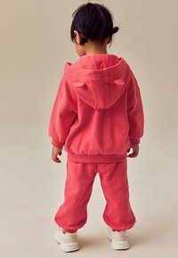 Lyserødt fleece-hættetrøje- og buksetøjssæt med ørneformede detaljer på hætten, med en afslappet pasform og ribkantede manchetter. Hvide sneakers fuldender looket.