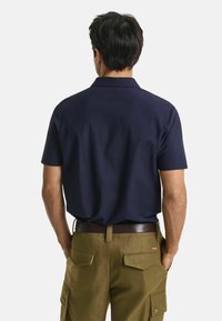 Chemise bleu marine à manches courtes avec un col, associée à un pantalon cargo beige avec des poches arrière plaquées et une ceinture marron. Tissu au toucher lisse.