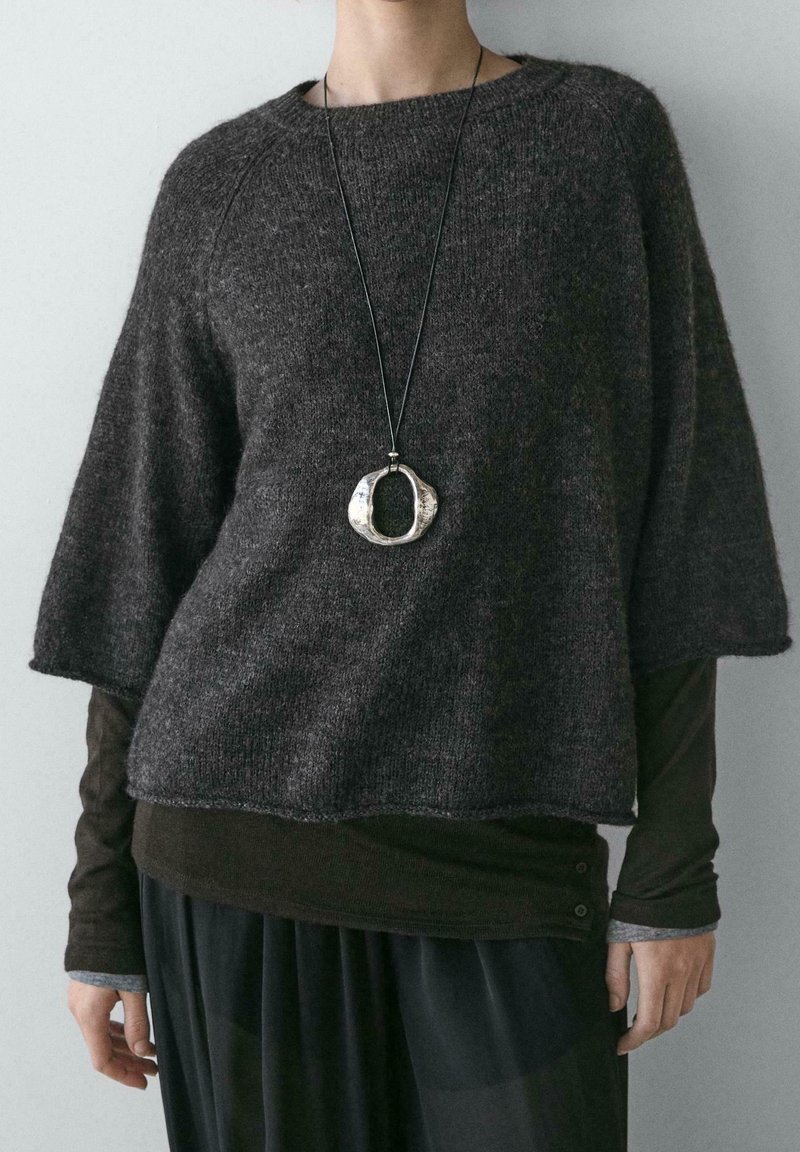 Personne portant un pull en maille texturée gris foncé à manches trois quarts, superposé à une chemise noire à manches longues, et un collier pendentif ovale argenté.