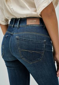 Jeans de mezclilla azul oscuro con un diseño ajustado, que cuentan con bolsillos traseros con costuras decorativas y una etiqueta de marca de cuero marrón en la cintura.