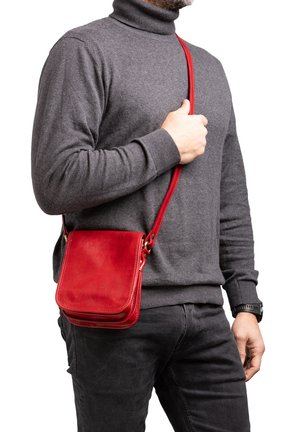 Man met een grijze coltrui, zwarte broek, zwart horloge, en een rode leren crossbodytas met klep.