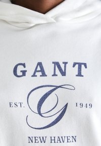 Bílá mikina s kapucí se námořnickým potiskem. Design zahrnuje "GANT," "EST. 1949" a "NEW HAVEN" v různých stylech písma. Měkká textilie.