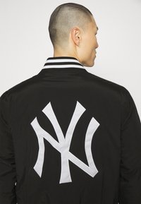 Chaqueta bomber negra con cuello de rayas blancas y gran logo blanco de NY en la parte posterior. Textura suave y diseño casual.