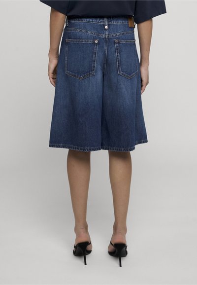 Personne portant un short en jean bleu foncé jusqu’aux genoux et des sandales à talons noirs, vue de dos sur un fond uni.