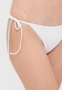 Biały dół bikini z regulowanymi sznureczkami po bokach, gładką strukturą i minimalnym przykryciem. Posiada klasyczny krój i czyste linie.