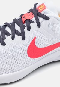 Hvite atletiske sko med teksturert mesh-overdel, grå lisser og en fremtredende rød Nike swoosh. Har polstret krage og oransje detaljer.