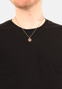 Roséguldhalsband med en cirkulär medaljong med en skorpiondesign, bärs över en svart T-shirt med rund halsringning.