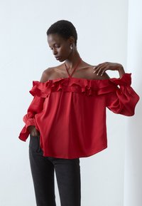 Blouse rouge sans manches avec des détails en volants, fabriquée à partir d'un tissu léger. Caractéristiques : manches longues et coupe décontractée. Associée à un pantalon noir.