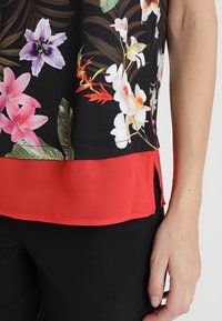 Top con motivo floreale su sfondo nero, caratterizzato da fiori multicolori vivaci e orlo rosso solido. Il tessuto sembra leggero e liscio.