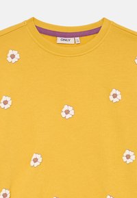 Felpa gialla con ricami floreali bianchi e dettagli rosa. Collo rotondo con un colletto interno viola a contrasto.
