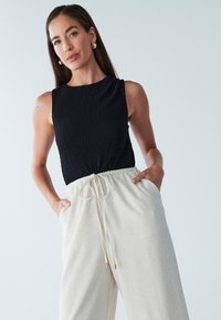 Melnā, teksturētā bezroku topa kombinācija ar gaiši bēšām biksēm ar aukliņu jostasvietā, kabatām un zeltainiem akcentiem.