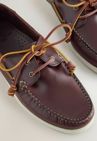 Chaussures bateau en cuir marron avec des lacets en cuir bordés de jaune noués en nœuds, semelles en caoutchouc blanches et coutures visibles le long des bords.