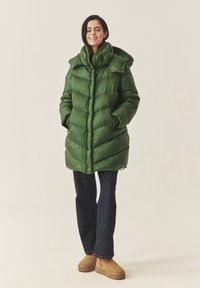 Cappotto gonfiato verde con colletto alto, design a righe orizzontali e chiusura con bottoni. Indossato sopra jeans a zampa d'elefante e stivaletti alla caviglia color beige.