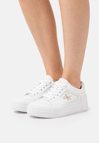 White Calvin Klein low-top sneakers with thick soles worn on feet against a plain light background.