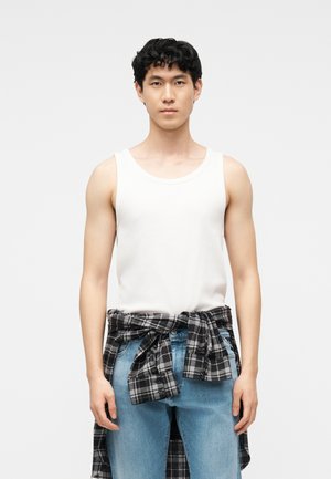 Joven con cabello negro y rizado que lleva una camiseta sin mangas blanca, jeans azul claro y una camisa de cuadros en blanco y negro atada alrededor de su cintura.