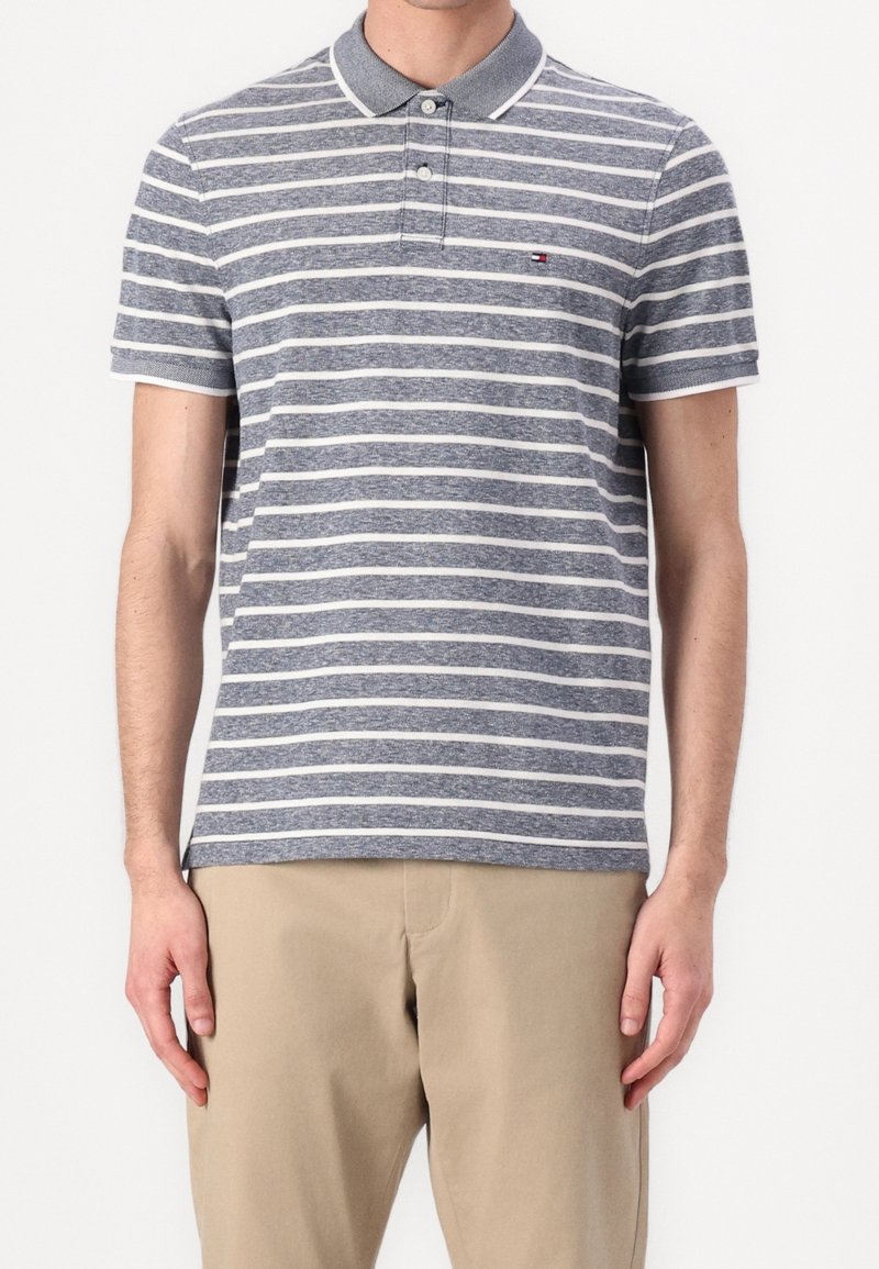 Homme portant un polo à rayures horizontales grises et blanches avec un pantalon beige, debout devant un fond blanc uni.