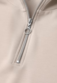 Gros plan d'un tissu beige clair avec une fermeture éclair métallique argentée partiellement ouverte, avec une tirette ronde sur le curseur.