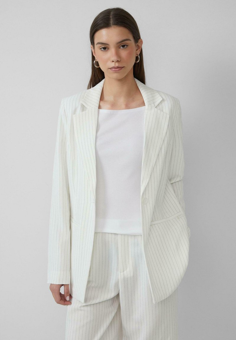 Femme avec de longs cheveux noirs portant un blazer à rayures blanches et un pantalon assorti par-dessus un haut blanc, se tenant contre un fond gris clair.